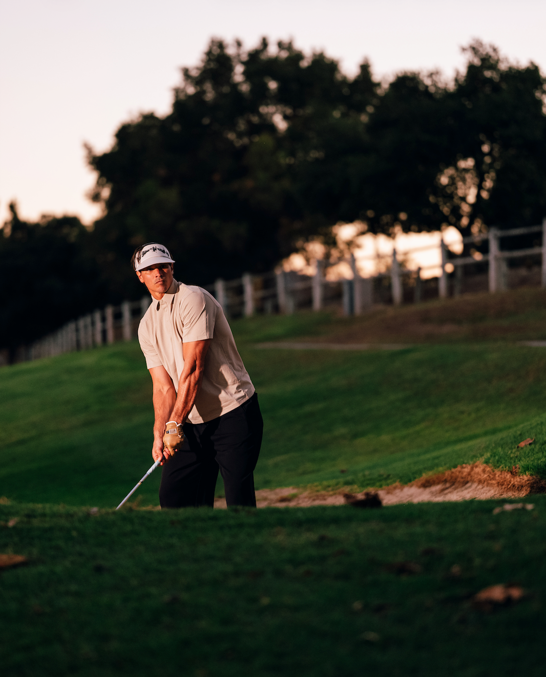Man playing golf on a green course with trees and a fence in the background.