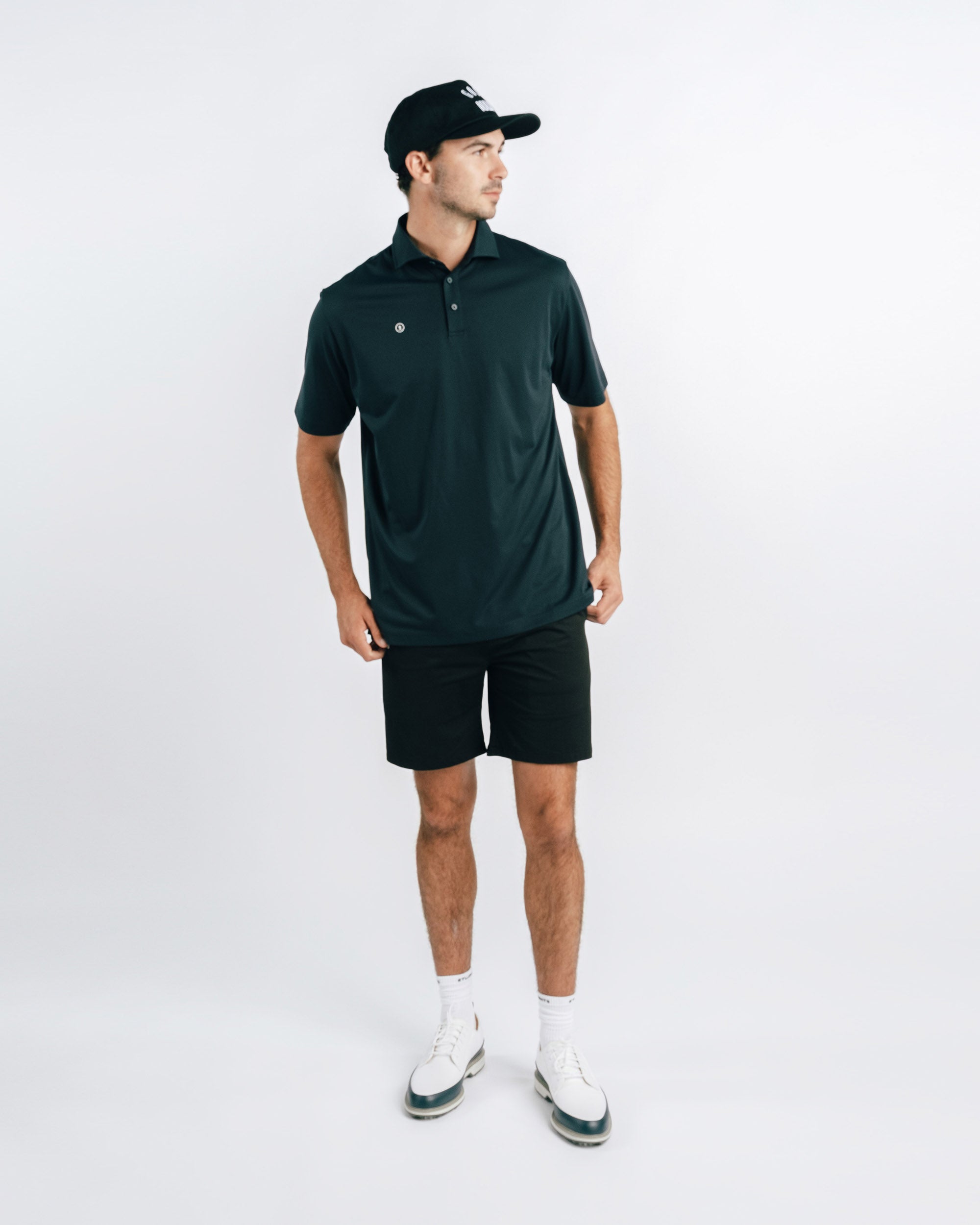 Man wearing a dark green polo shirt, black shorts, and white shoes on a white background