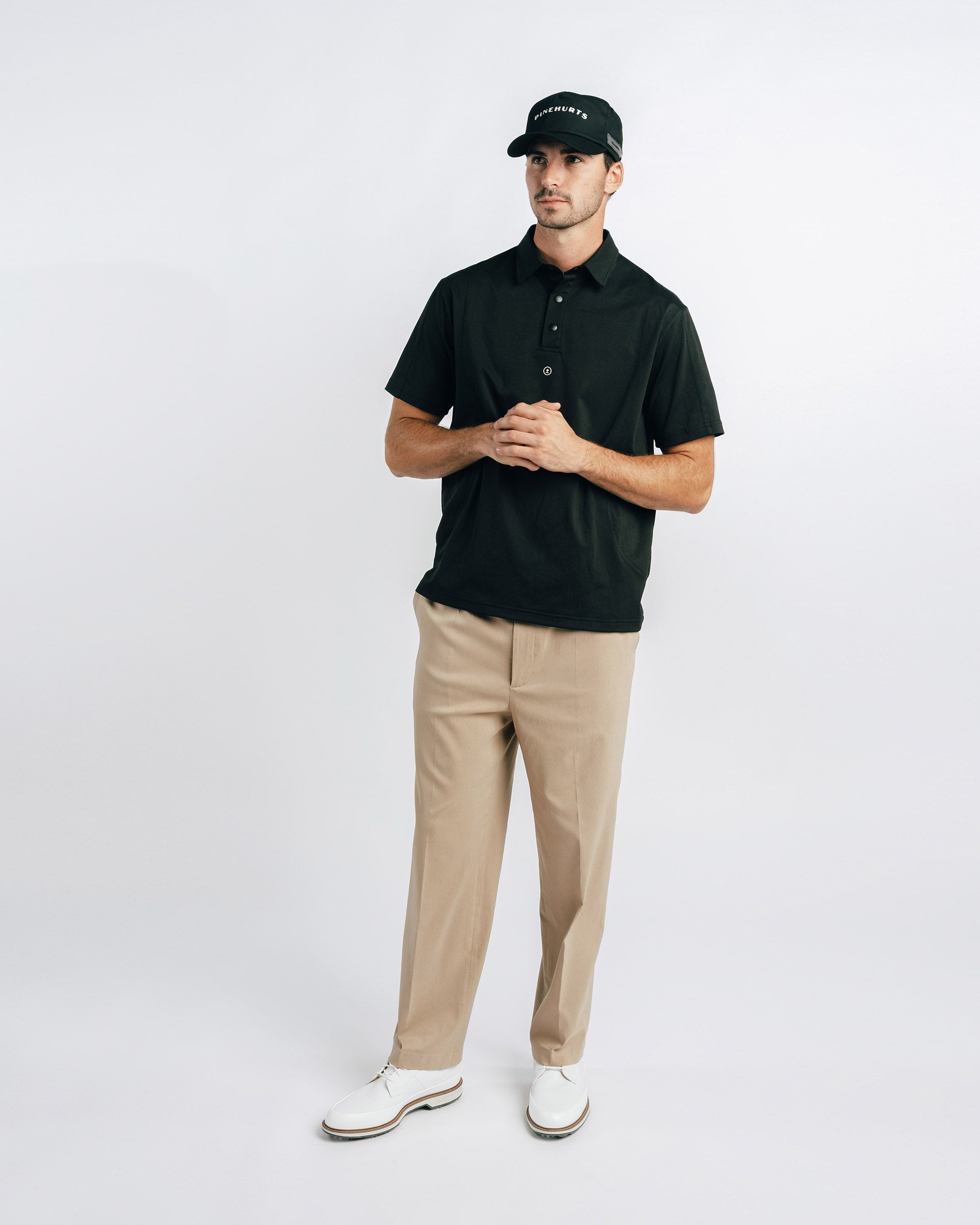 Man wearing a black polo shirt, beige pants, and white shoes on a white background