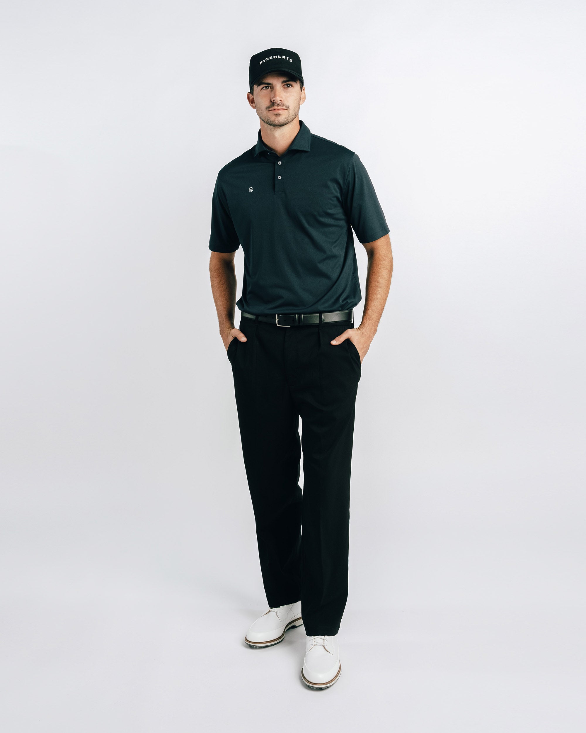 Man wearing a green polo shirt, black pants, and a black cap on a white background