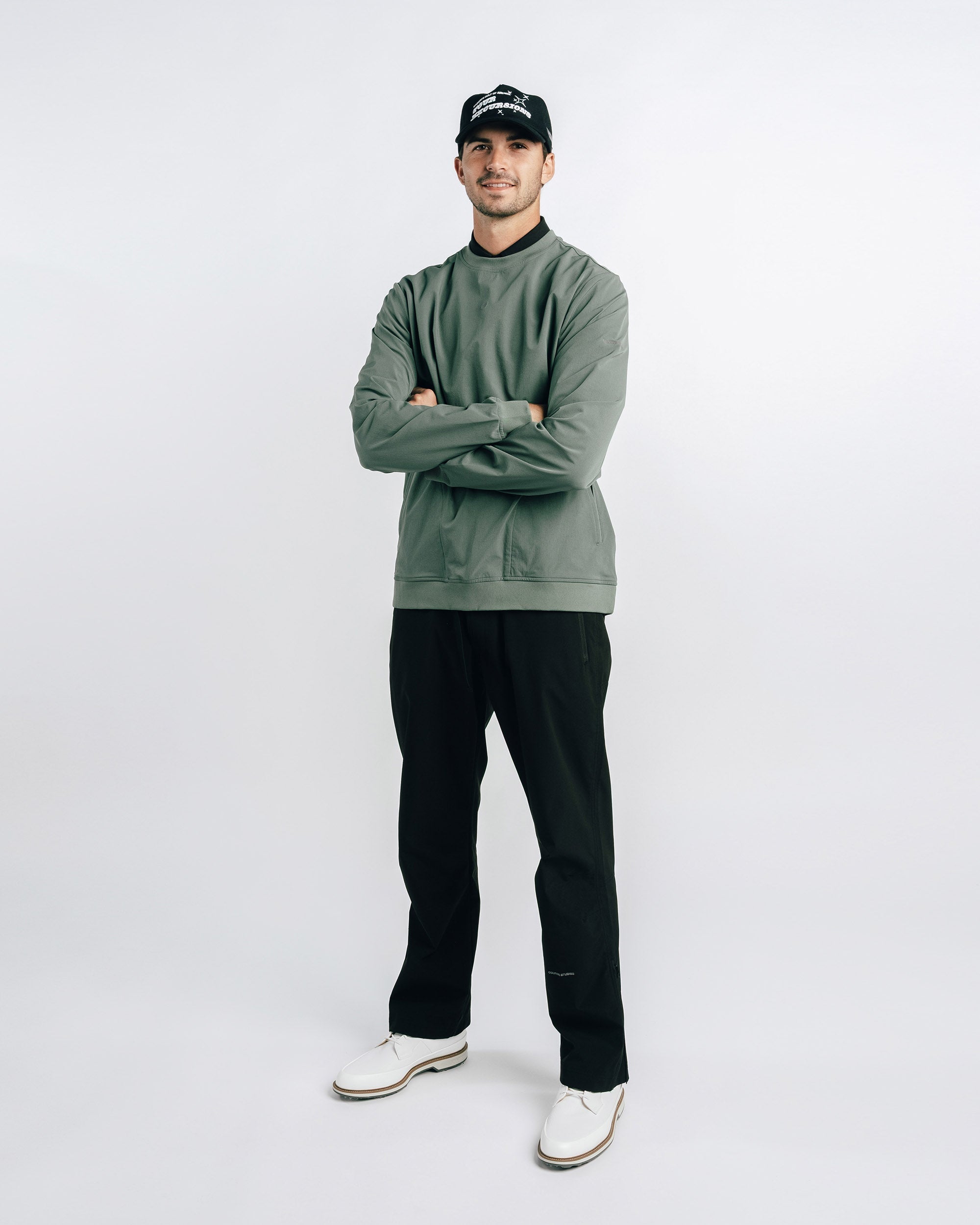 Man wearing a green sweater, black pants, and a black cap on a white background
