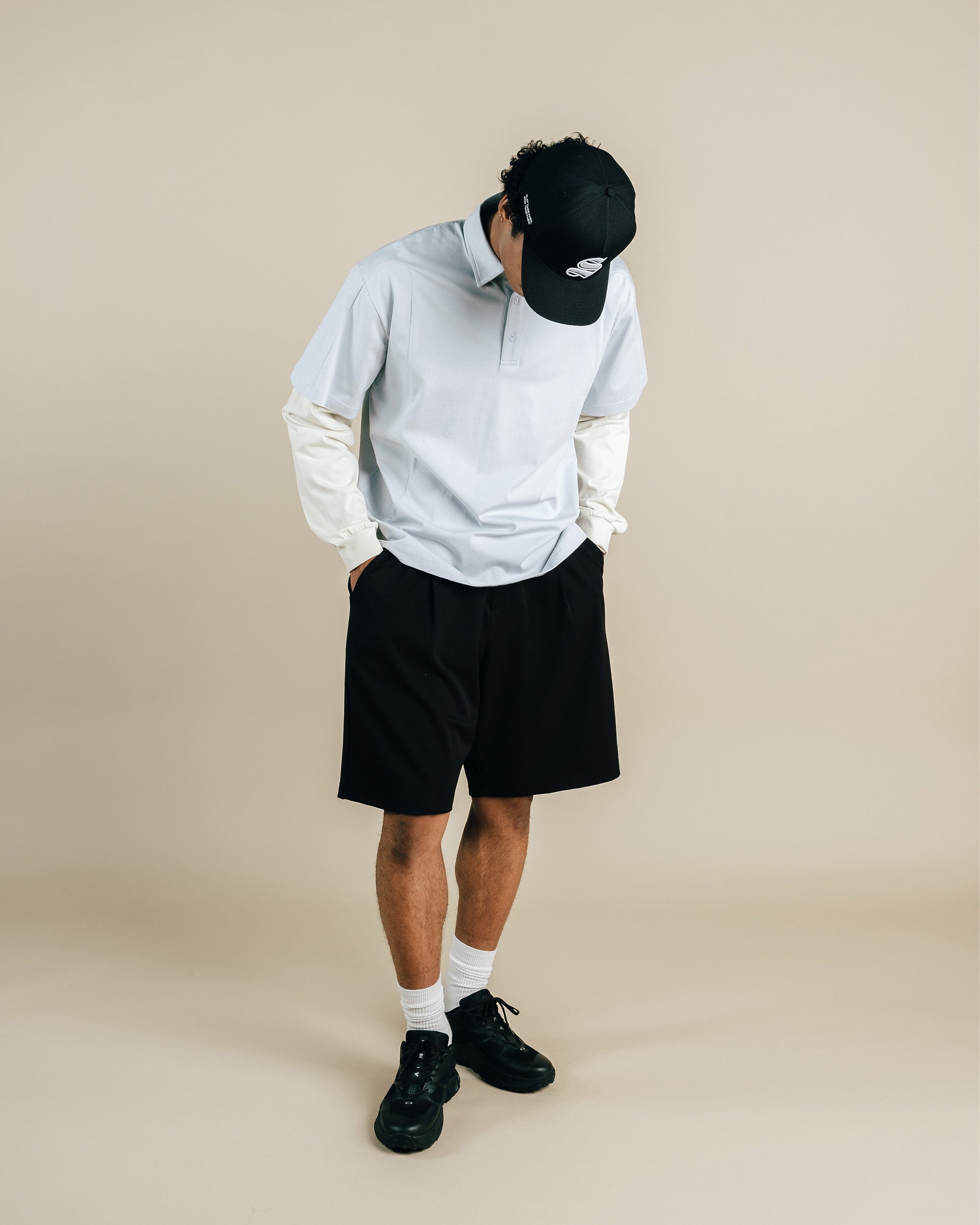 Person wearing a light gray hoodie, black shorts, and black cap on a beige background