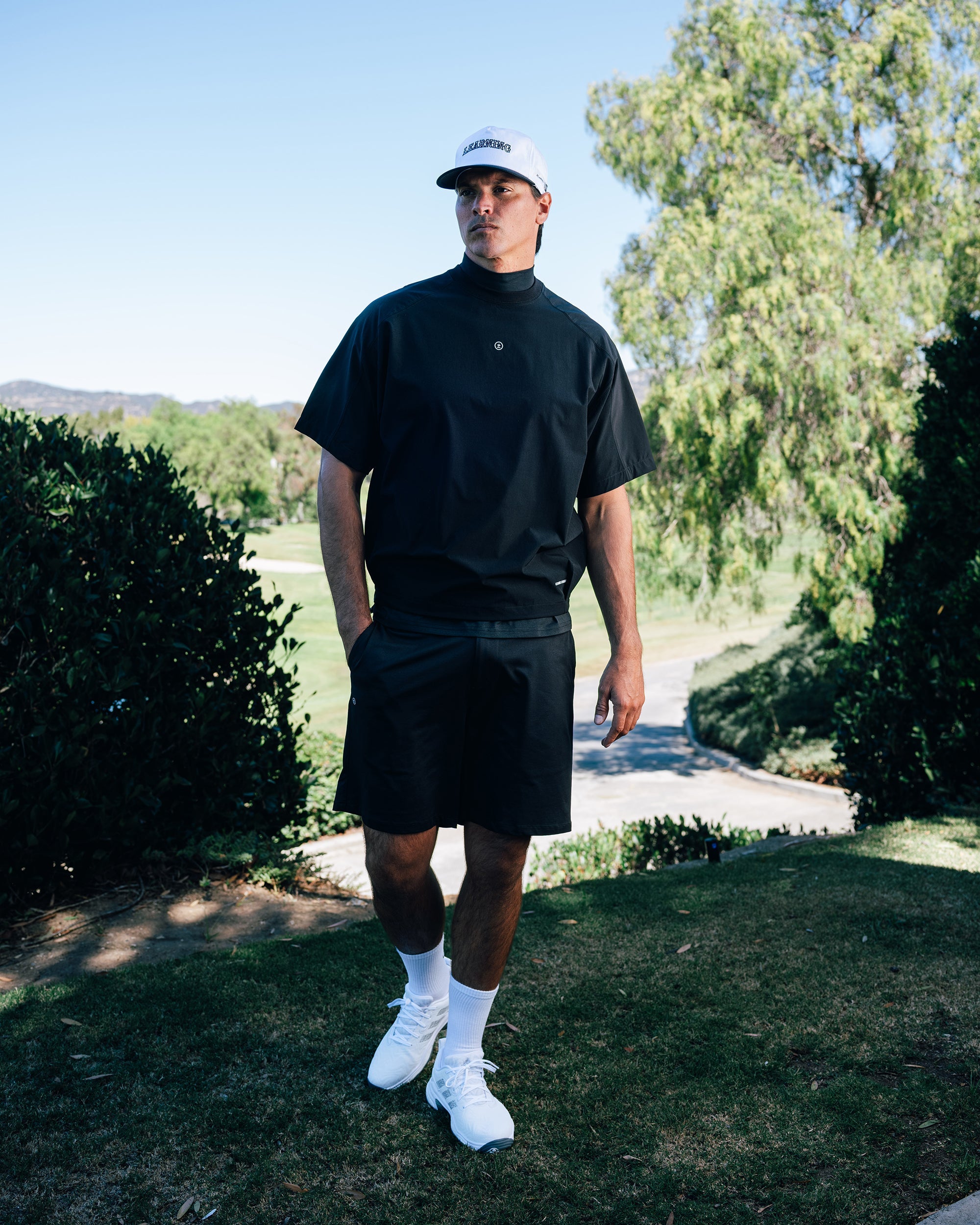 Person wearing a black outfit with white shoes standing outdoors on a sunny day.