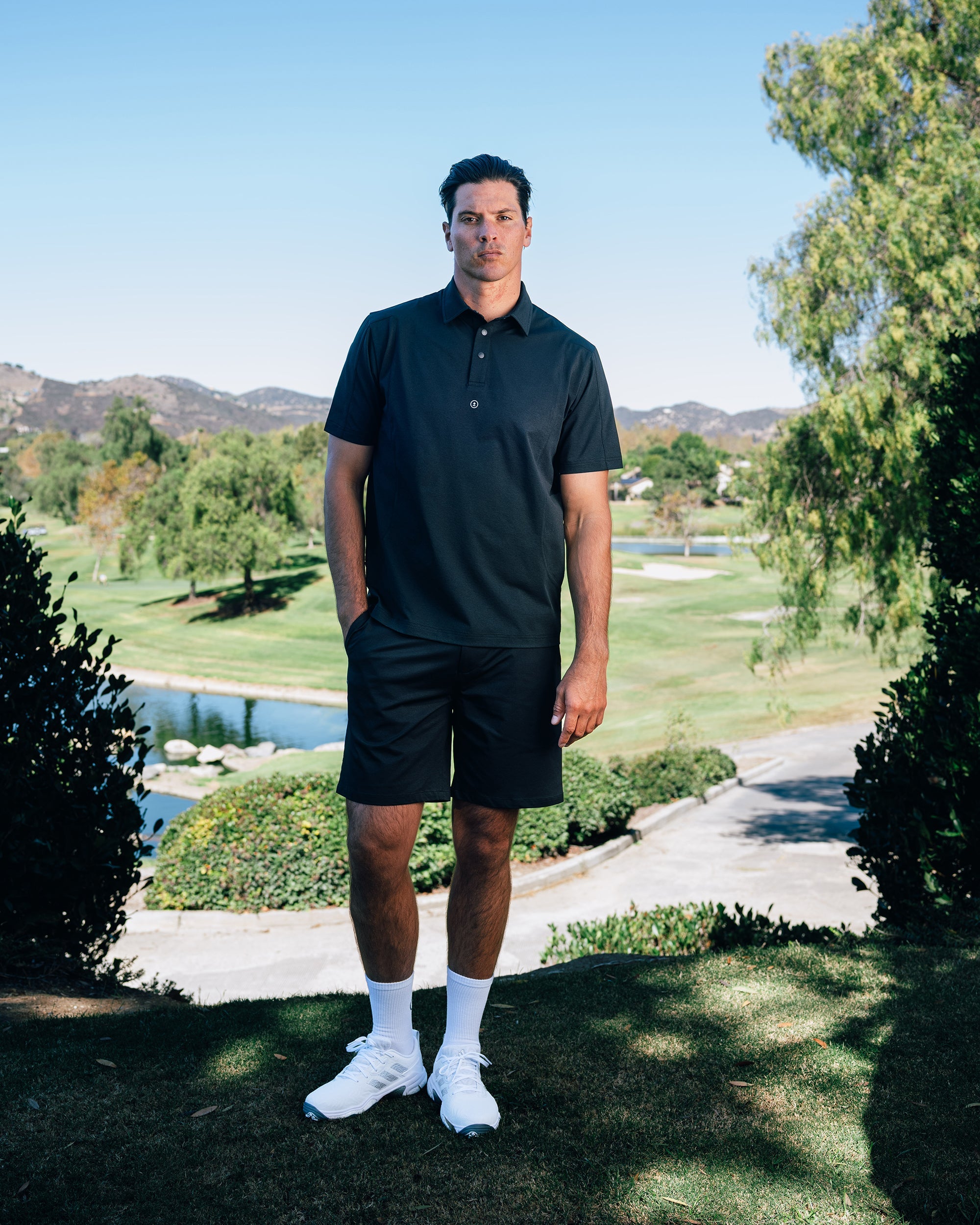Man in black polo shirt and shorts standing on a golf course with mountains in the background