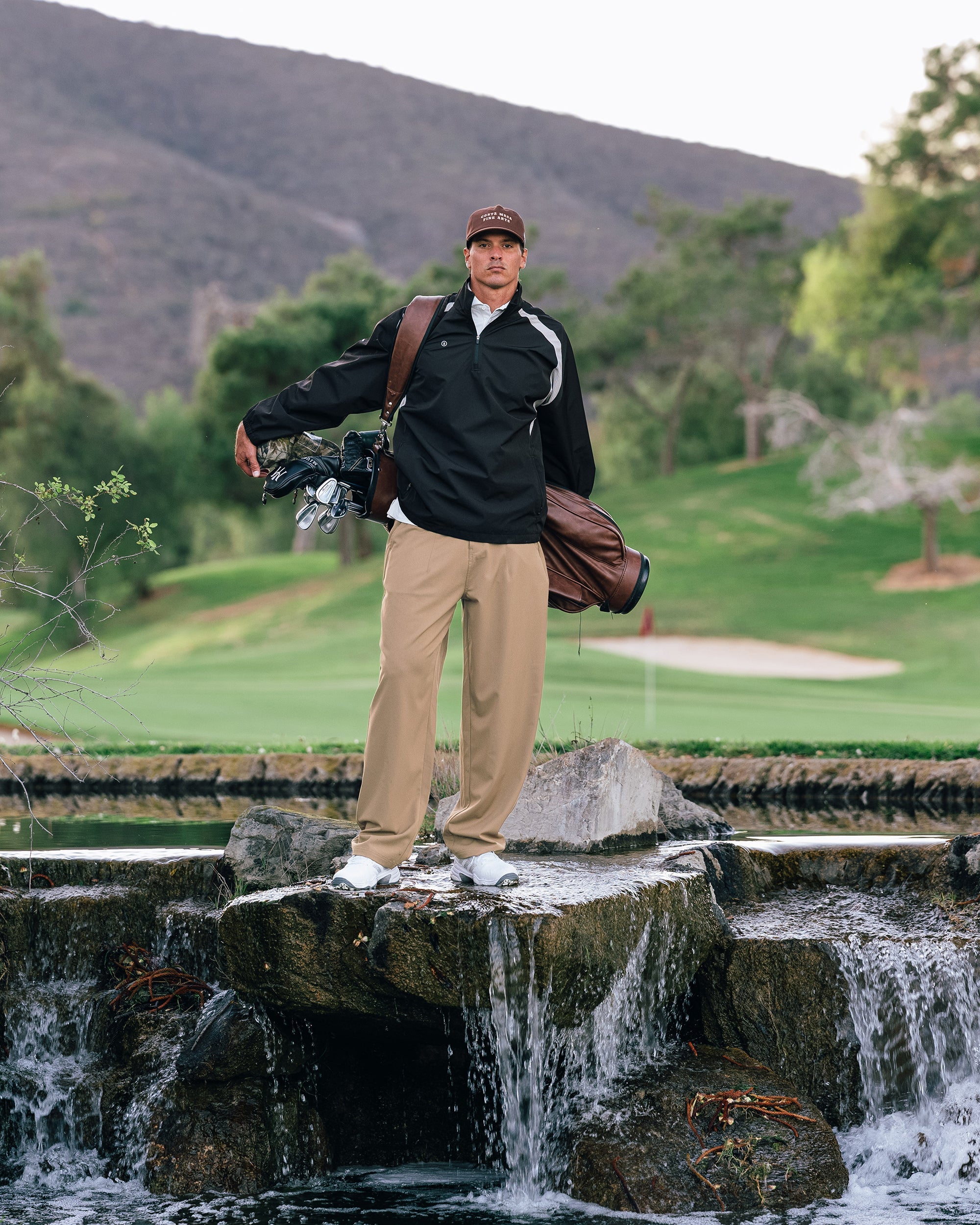 Man standing on a stone ledge with a golf club and bag in a golf course setting