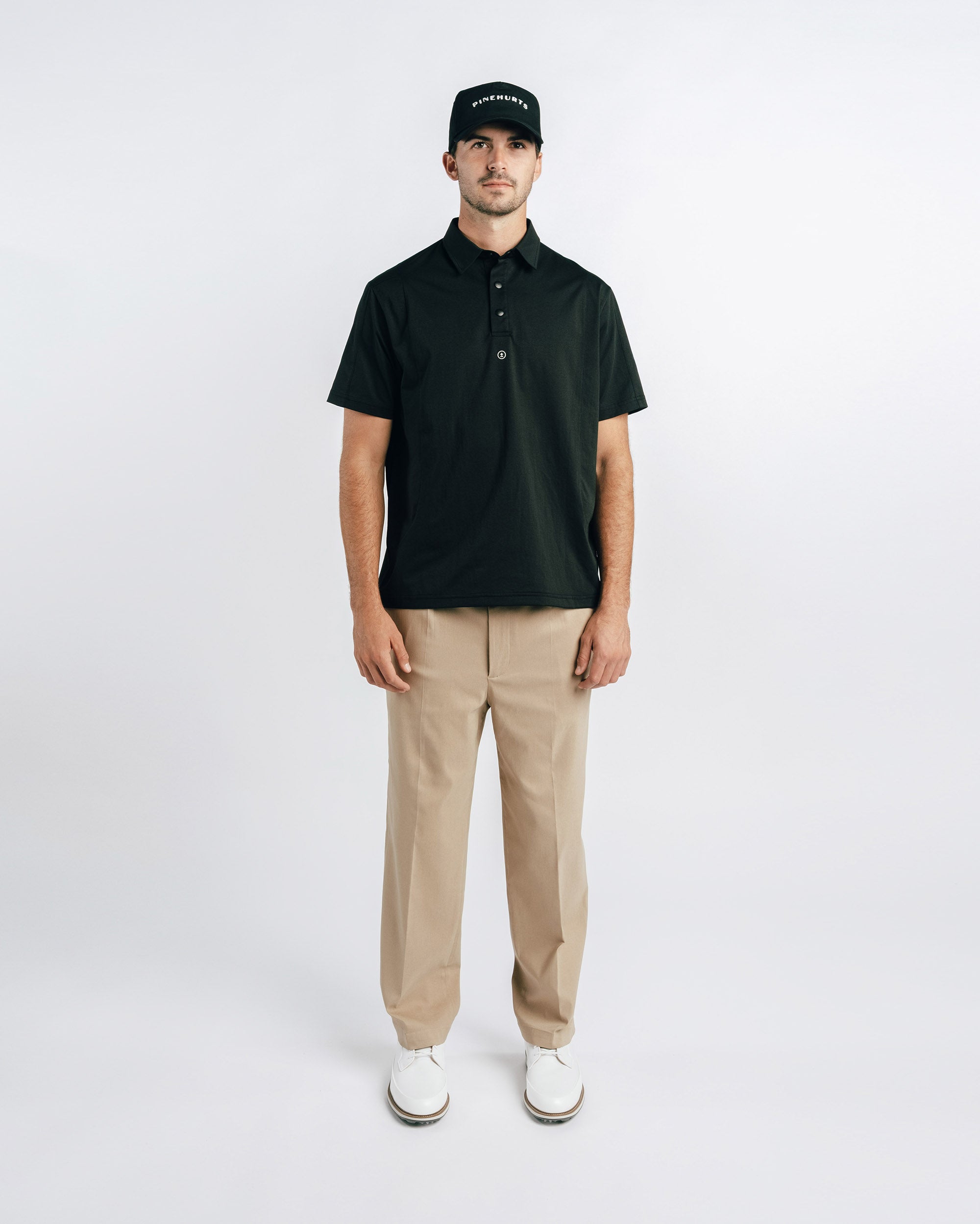 Man wearing a black polo shirt, beige pants, and white shoes on a white background