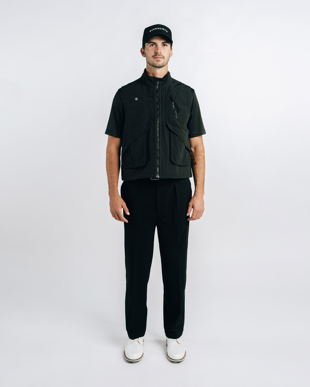 Man wearing a black vest, black shirt, black pants, and white shoes on a white background