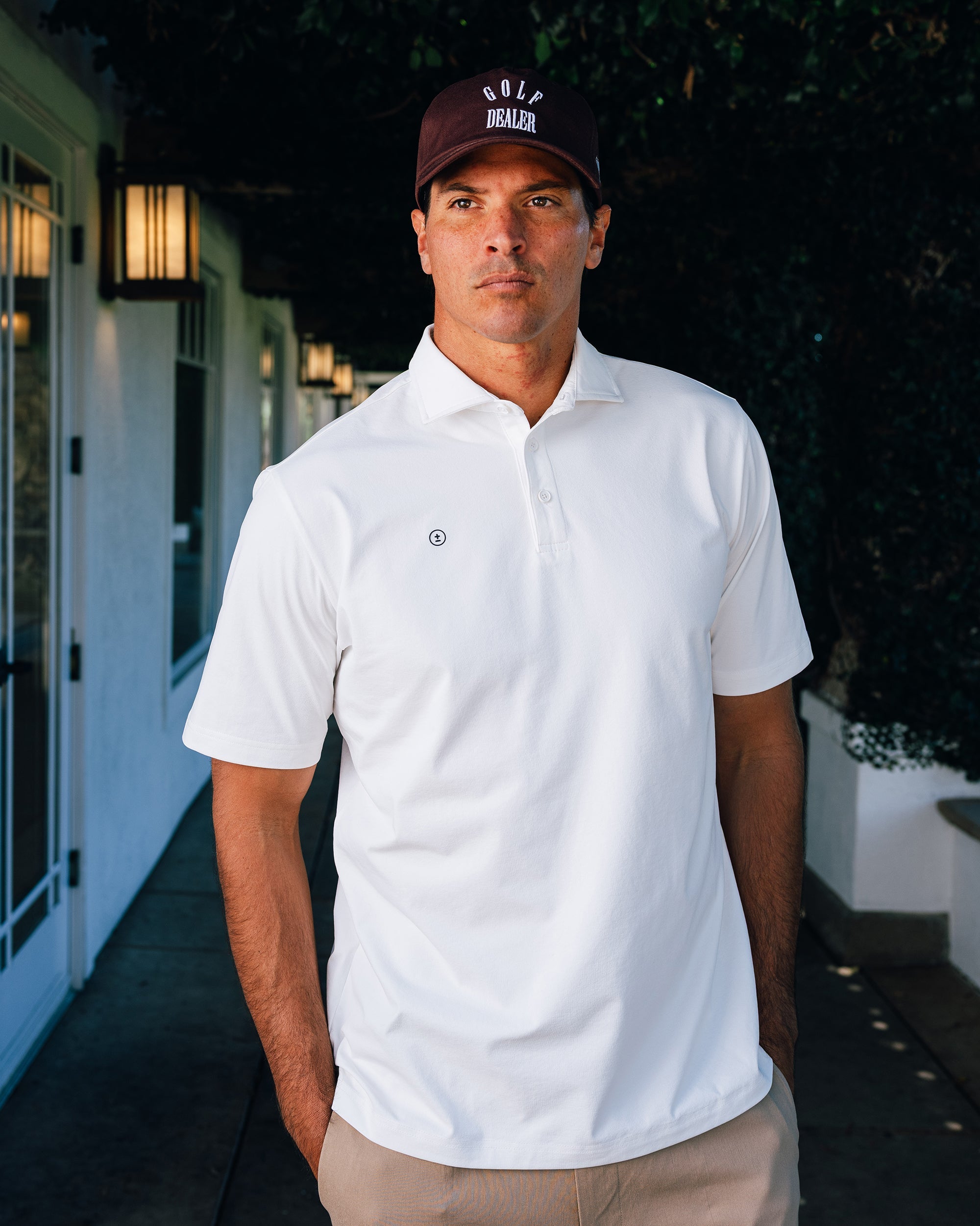 Man wearing a white polo shirt and brown cap standing outdoors.