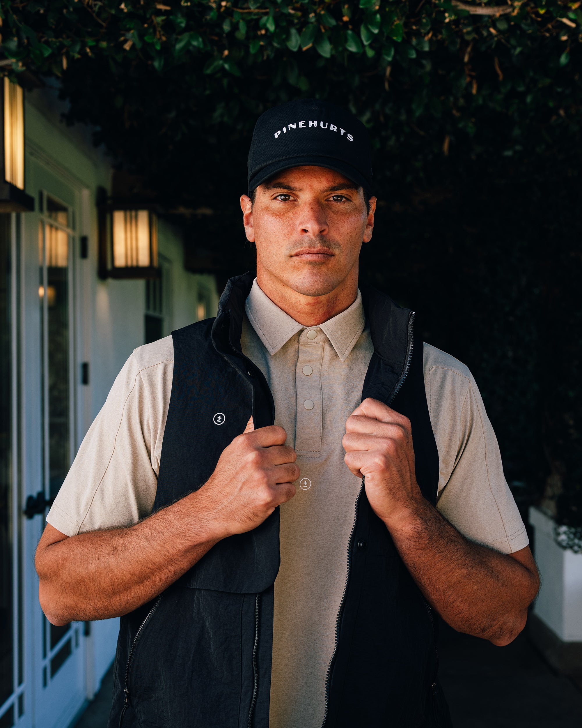 Man wearing a black cap and vest standing outdoors with greenery in the background