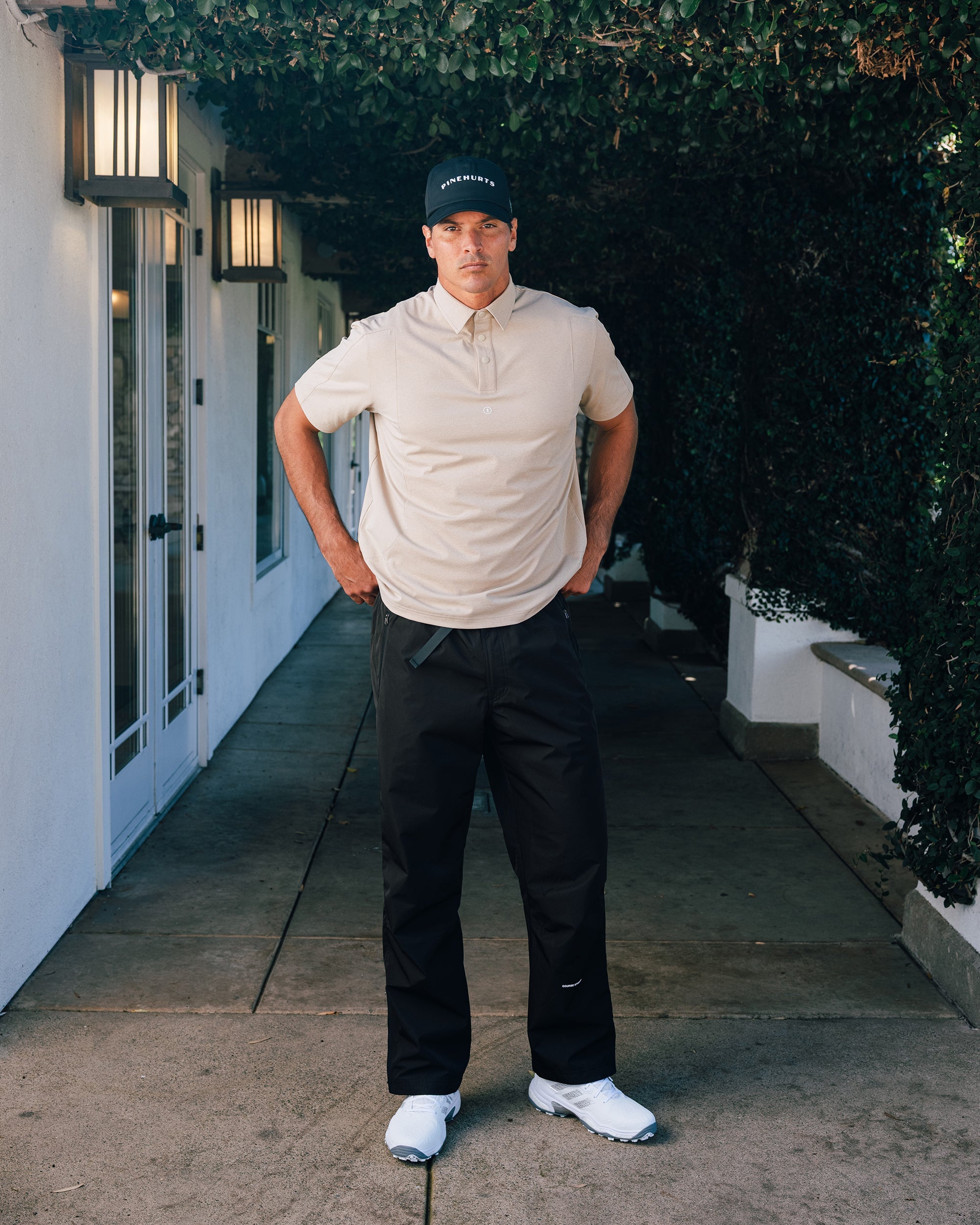 Man in golf attire standing on a path with greenery and a building in the background