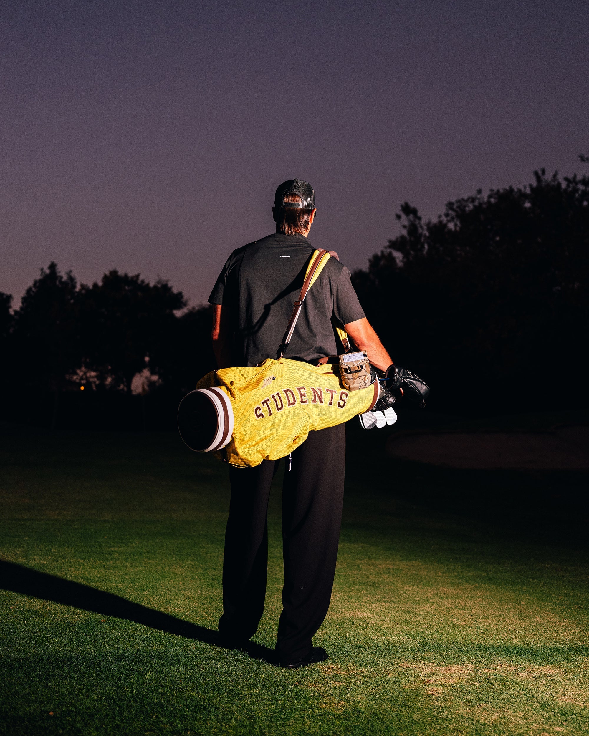 Person walking on a golf course with a yellow bag labeled 'Students' and golf clubs.
